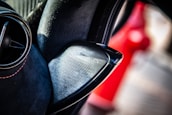 A close-up of a car's speaker system, focusing on the Bowers & Wilkins speaker integrated into a plush black interior with red stitching. A ventilation grille is visible to the left, and there is a blurred red object in the background suggesting a vibrant and modern car interior.