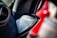 A close-up of a car's speaker system, focusing on the Bowers & Wilkins speaker integrated into a plush black interior with red stitching. A ventilation grille is visible to the left, and there is a blurred red object in the background suggesting a vibrant and modern car interior.