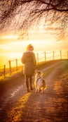 woman in brown coat holding white dog on brown dirt road during sunset