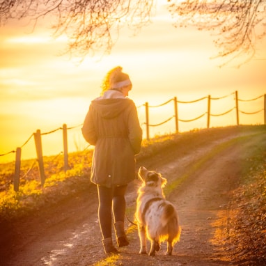 woman in brown coat holding white dog on brown dirt road during sunset