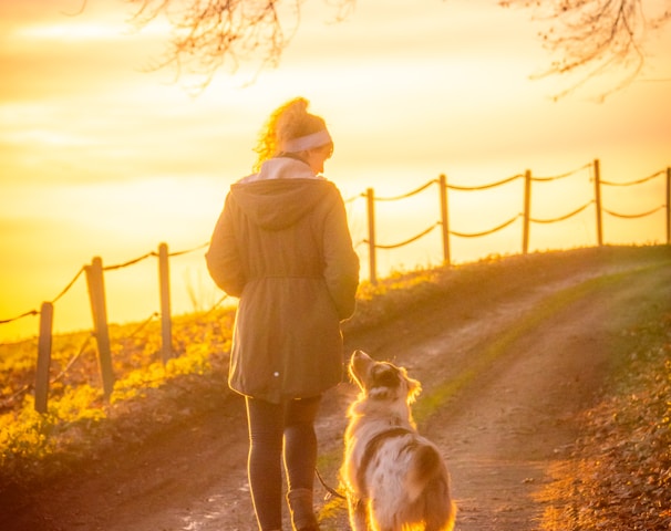 woman in brown coat holding white dog on brown dirt road during sunset