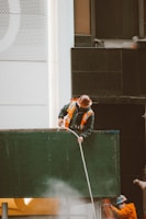 Two construction workers wearing hard hats and reflective vests are engaged in work on a building site. One worker is standing on top of a green structure, holding a rope, while the other worker is partially visible in the background, wearing an orange uniform. The building behind them has a modern facade with a mix of white and dark panels and perforated designs.