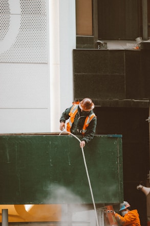 Two construction workers wearing hard hats and reflective vests are engaged in work on a building site. One worker is standing on top of a green structure, holding a rope, while the other worker is partially visible in the background, wearing an orange uniform. The building behind them has a modern facade with a mix of white and dark panels and perforated designs.