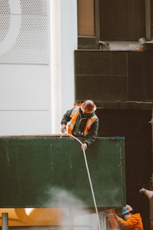 Two construction workers wearing hard hats and reflective vests are engaged in work on a building site. One worker is standing on top of a green structure, holding a rope, while the other worker is partially visible in the background, wearing an orange uniform. The building behind them has a modern facade with a mix of white and dark panels and perforated designs.
