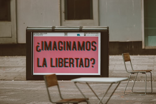 A public space features a large sign with bold, colorful text that reads '¿Imaginamos la libertad?' in Spanish, suggesting a thought-provoking question about freedom. Surrounding the sign are a few empty chairs and tables, indicating an outdoor seating area. The background consists of gray urban architecture.