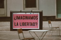 A public space features a large sign with bold, colorful text that reads '&iquest;Imaginamos la libertad?' in Spanish, suggesting a thought-provoking question about freedom. Surrounding the sign are a few empty chairs and tables, indicating an outdoor seating area. The background consists of gray urban architecture.
