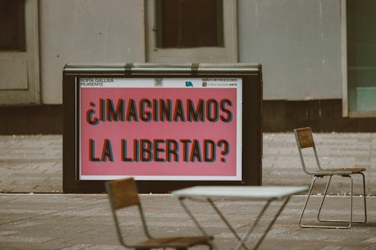 A public space features a large sign with bold, colorful text that reads '&iquest;Imaginamos la libertad?' in Spanish, suggesting a thought-provoking question about freedom. Surrounding the sign are a few empty chairs and tables, indicating an outdoor seating area. The background consists of gray urban architecture.