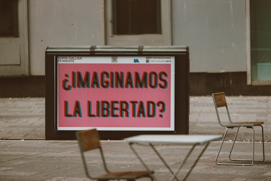 A public space features a large sign with bold, colorful text that reads '&iquest;Imaginamos la libertad?' in Spanish, suggesting a thought-provoking question about freedom. Surrounding the sign are a few empty chairs and tables, indicating an outdoor seating area. The background consists of gray urban architecture.