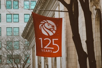 A red flag displaying a stylized white lion head logo with the text '125 Years' is hanging on the side of a classic architectural building. The surroundings feature a few bare trees, and a nearby modern building with a grid of turquoise windows is visible in the background.