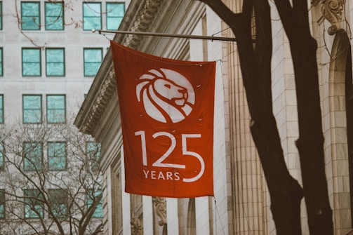 A red flag displaying a stylized white lion head logo with the text '125 Years' is hanging on the side of a classic architectural building. The surroundings feature a few bare trees, and a nearby modern building with a grid of turquoise windows is visible in the background.
