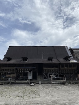 A large, traditional building with a dark corrugated metal roof and open eaves. The structure appears to be wooden with various sections and spaces underneath the eaves. There is a paved courtyard in front of the building and a mix of objects such as a scaffold, stacked materials, and a freestanding door frame resting against the building.