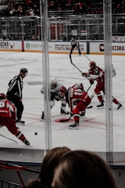Players on an ice hockey rink are engaged in a faceoff. Two teams, dressed in red and white uniforms, compete intensely as referees oversee the action. The scene is energetic, with the ice covered by hockey markings and surrounded by protective glass barriers. Spectators are visible in the background.