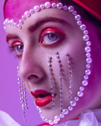 A close-up portrait showcases intricate makeup artistry, featuring vibrant red and pink eyeshadow, and striking red lipstick. Pearls and crystals delicately embellish the face, creating a theatrical and avant-garde style against a purple background.