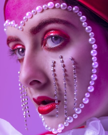 A close-up portrait showcases intricate makeup artistry, featuring vibrant red and pink eyeshadow, and striking red lipstick. Pearls and crystals delicately embellish the face, creating a theatrical and avant-garde style against a purple background.