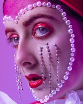 A close-up portrait showcases intricate makeup artistry, featuring vibrant red and pink eyeshadow, and striking red lipstick. Pearls and crystals delicately embellish the face, creating a theatrical and avant-garde style against a purple background.