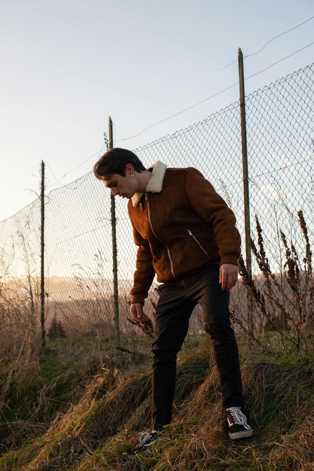 man in brown jacket and black pants standing beside gray metal fence during daytime