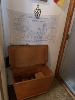 A wooden chest with an open lid is situated beneath a decorative wall hanging. The hanging features a pastoral scene with trees, a shepherd, and sheep, accompanied by text. Above the hanging, a wrought iron decorative piece is mounted on the wall. A flag is partially visible in the corner of the image.