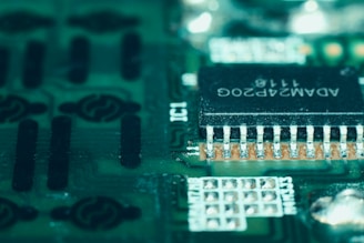 A close-up view of a green circuit board featuring a prominent black microchip with metallic pins. The microchip has white alphanumeric markings on its surface, and the surrounding area displays various circuit patterns and solder points.