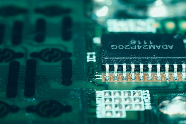 A close-up view of a green circuit board featuring a prominent black microchip with metallic pins. The microchip has white alphanumeric markings on its surface, and the surrounding area displays various circuit patterns and solder points.