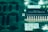 A close-up view of a green circuit board featuring a prominent black microchip with metallic pins. The microchip has white alphanumeric markings on its surface, and the surrounding area displays various circuit patterns and solder points.
