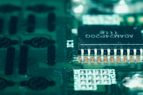 A close-up view of a green circuit board featuring a prominent black microchip with metallic pins. The microchip has white alphanumeric markings on its surface, and the surrounding area displays various circuit patterns and solder points.