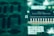 A close-up view of a green circuit board featuring a prominent black microchip with metallic pins. The microchip has white alphanumeric markings on its surface, and the surrounding area displays various circuit patterns and solder points.
