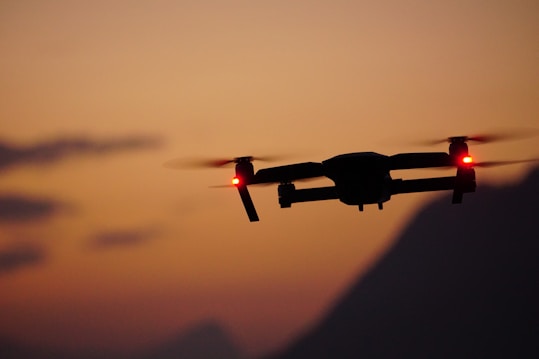 A silhouette of a drone with red lights on its wings flying against a sunset backdrop, with a gradient sky transitioning from orange to dusky blue. The scene includes blurred mountain outlines in the background, adding depth to the composition.