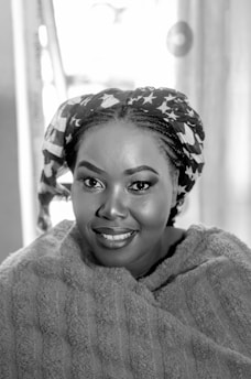 A woman with a patterned headscarf and a textured wrap smiles warmly. The background is softly blurred, drawing focus to her expressive eyes and subtle makeup, providing a sense of elegance and poise.