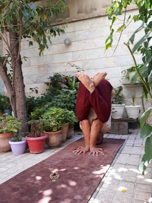 A person is practicing a yoga pose on a brown mat in a serene garden setting, surrounded by various potted plants and trees. The individual is inverted, balancing on their forearms with legs bent over their head. The stone wall in the background and green foliage create a peaceful atmosphere.