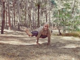A person is performing a yoga or acrobatic pose in the middle of a forest, balancing on their hands with a serene and focused expression. The forest is dense with tall trees and dappled sunlight filtering through the leaves.