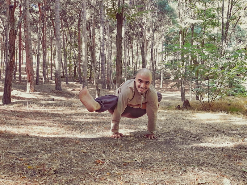 A person is performing a yoga or acrobatic pose in the middle of a forest, balancing on their hands with a serene and focused expression. The forest is dense with tall trees and dappled sunlight filtering through the leaves.