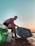A person with a fit physique is sitting on the edge of a boat, holding a striped wakeboard. The setting appears to be during sunset or sunrise, with a calm body of water surrounding the boat. The sky transitions from a soft blue at the top to warm orange hues near the horizon.