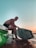 A person with a fit physique is sitting on the edge of a boat, holding a striped wakeboard. The setting appears to be during sunset or sunrise, with a calm body of water surrounding the boat. The sky transitions from a soft blue at the top to warm orange hues near the horizon.