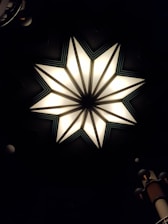 A geometric star-shaped light fixture with eight points, featuring bright illumination and contrasting dark shadowed areas. The star design includes triangular segments with soft white light, bordered by darker lines that create a striking focal point against a dim background. Peripheral objects include segments of cylindrical structures with vertical ridges.