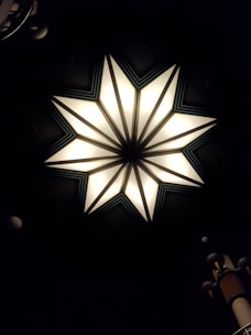 A geometric star-shaped light fixture with eight points, featuring bright illumination and contrasting dark shadowed areas. The star design includes triangular segments with soft white light, bordered by darker lines that create a striking focal point against a dim background. Peripheral objects include segments of cylindrical structures with vertical ridges.