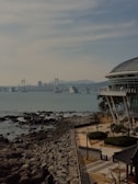 A coastal scene featuring a modern building with a curved roof, located near the water. In the background, a large suspension bridge spans across the water with a cityscape and mountains visible. The foreground includes rocks along the shoreline and a landscaped area with trees and walking paths.