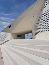 A modern architectural structure with geometric patterns, featuring white concrete steps leading up to a large building with a distinctive angular roof. The surface is covered with a combination of smooth and textured panels, and small windows are scattered across the facade.