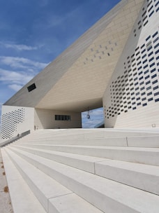 A modern architectural structure with geometric patterns, featuring white concrete steps leading up to a large building with a distinctive angular roof. The surface is covered with a combination of smooth and textured panels, and small windows are scattered across the facade.