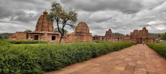 An ancient temple complex with intricate stone architecture set against a cloudy sky. The foreground features a neatly trimmed green hedge lining a paved pathway, leading to the temple structures. The temple buildings are made of sandstone, exhibiting detailed carvings and South Indian architectural elements.