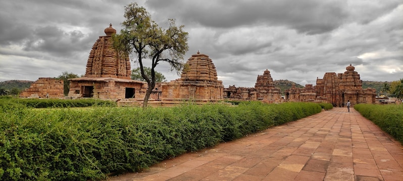 An ancient temple complex with intricate stone architecture set against a cloudy sky. The foreground features a neatly trimmed green hedge lining a paved pathway, leading to the temple structures. The temple buildings are made of sandstone, exhibiting detailed carvings and South Indian architectural elements.