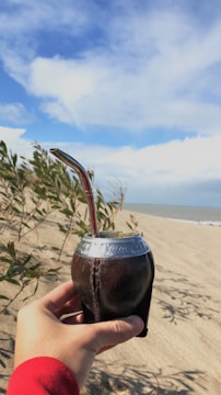 A hand holding a traditional mate gourd with a metal straw, set against a sandy beach backdrop and a bright blue sky with scattered clouds. Some green plants are visible in the foreground.