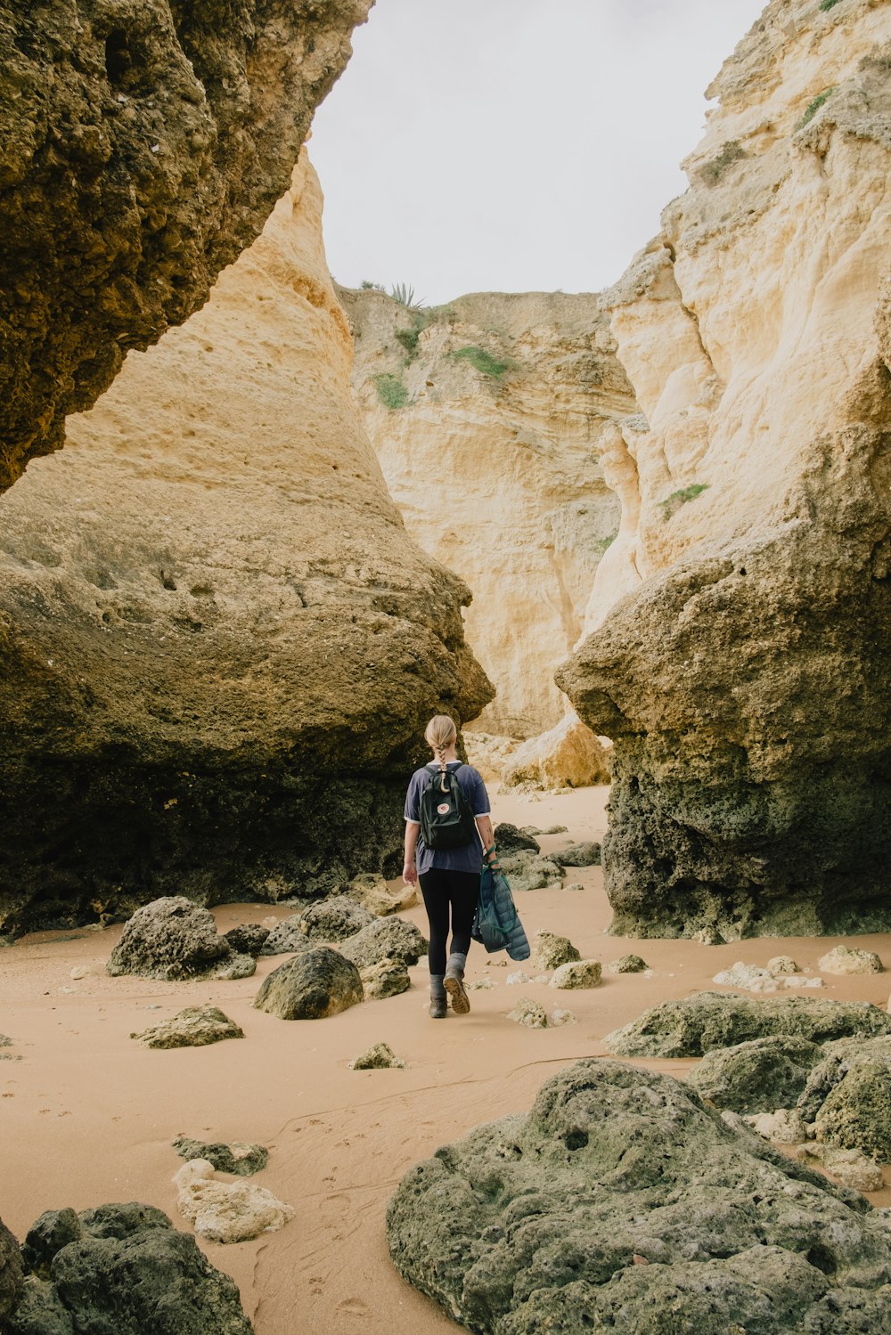 man in blue jacket and black pants standing on brown sand near brown rock formation during