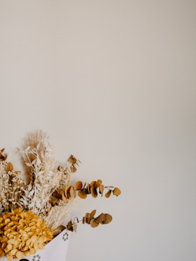 A dried floral arrangement featuring various plants with golden and brown hues, including eucalyptus and possibly hydrangeas. The background is a plain, light beige or cream color, providing a minimalist and rustic aesthetic.