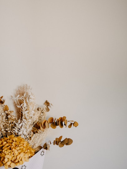 A dried floral arrangement featuring various plants with golden and brown hues, including eucalyptus and possibly hydrangeas. The background is a plain, light beige or cream color, providing a minimalist and rustic aesthetic.