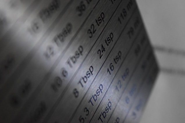 A close-up view of a metal conversion chart listing measurements in tablespoons and teaspoons alongside their metric equivalents. The surface has a brushed metallic finish, and the text is engraved or printed onto it.