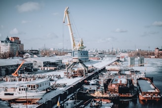 A busy shipyard scene featuring a large crane positioned on a dock, surrounded by various ships and industrial buildings. The area is covered in snow, indicating a cold winter environment. Surrounding the dock are several buildings and distant city structures, under a partly cloudy sky.
