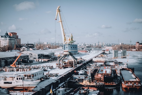 A busy shipyard scene featuring a large crane positioned on a dock, surrounded by various ships and industrial buildings. The area is covered in snow, indicating a cold winter environment. Surrounding the dock are several buildings and distant city structures, under a partly cloudy sky.