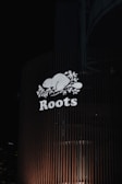 A brightly illuminated sign featuring the word 'Roots' with a stylized beaver and maple leaves. The sign is mounted on a building with vertical slats, set against a dark night sky. City lights are faintly visible in the background.