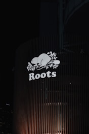A brightly illuminated sign featuring the word 'Roots' with a stylized beaver and maple leaves. The sign is mounted on a building with vertical slats, set against a dark night sky. City lights are faintly visible in the background.