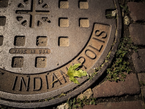 A manhole cover with the text 'INDIANAPOLIS' and 'MADE IN USA' is partially surrounded by green grass. A single green leaf is resting on the cover, adding a touch of nature to the otherwise industrial setting.